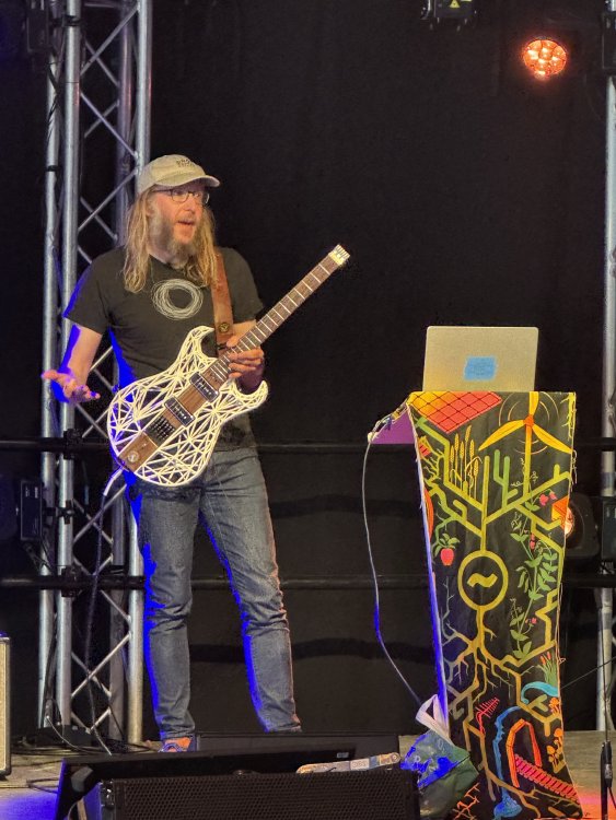 A photo of me on a stage behind a lectern, holding a weird guitar that is made from a wooden core with a 3D-printed body.