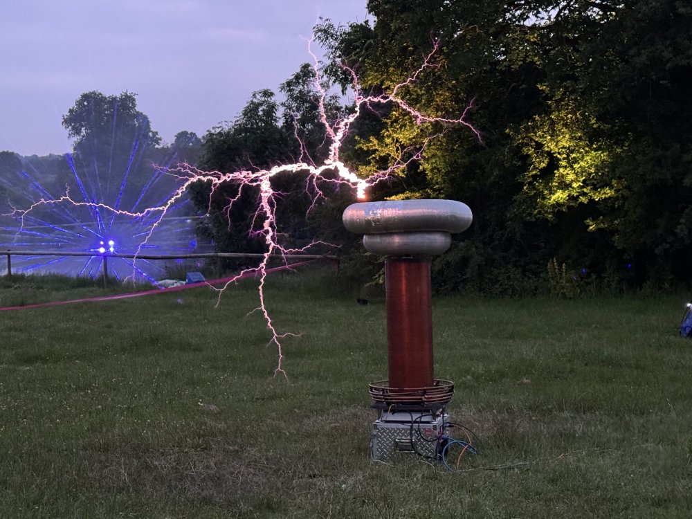 A photo of a tesla coil in a field at night doing its thing. The coil is a 2 metre high metal cylinder with a larger top section, and coming out of that for several metres is an arc of electricity lighting the area up.