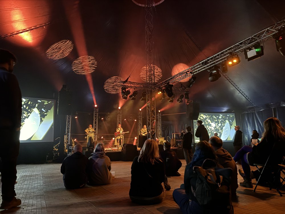 A photo of a group of people sat in a large event tent watching a stage, on which there are three performers: the man on the left has a laptop, the woman in the middle has a guitar and a mic stand into which she's signing, and the man on the right has a bass guitar. Either side of the stage are large screens with trippy computer graphics on.