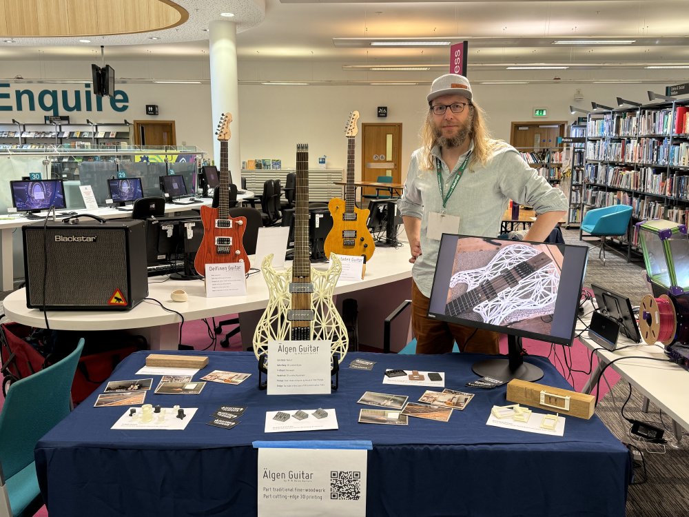 A photo of me stood in a library behind a table with several guitars, including one where the body is 3D printed.