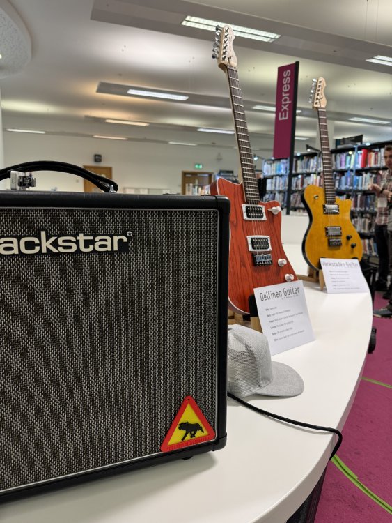 A photo of the area behind my table, showing a blackstar amp and two guitars, one an orange offset-style guitar called Delfinen, and the other a yellow guitar called Verkstaden. On the amp is stuck a small moose crossing sign as you might see in Sweden.