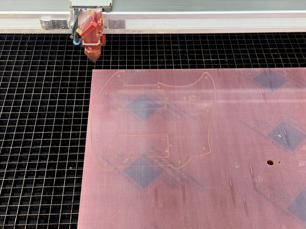 A photo of a sheet of vaguely orange acrylic with the protective film on, sat on the bed of a laser-cutter. Cut into the sheet is the familiar outline of a guitar pick-guard.