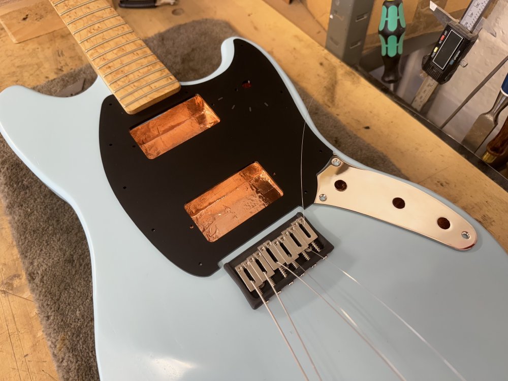 A photo of an offset-style electric guitar with a blue painted body and a birds-eye maple neck sitting on a workbench. There are not electronics installed, but there is now a matt-black pick-guard sat on it, along with a chrome metal piece where the volume, tone and input jack will all mount.