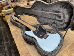 A photo of an electric guitar sat in a case on a workbench in a workshop. The guitar is an offset design, with a pale blue painted body, a black pickguard, with two chrome humbucker pickups on it. The neck is a birds-eye maple.