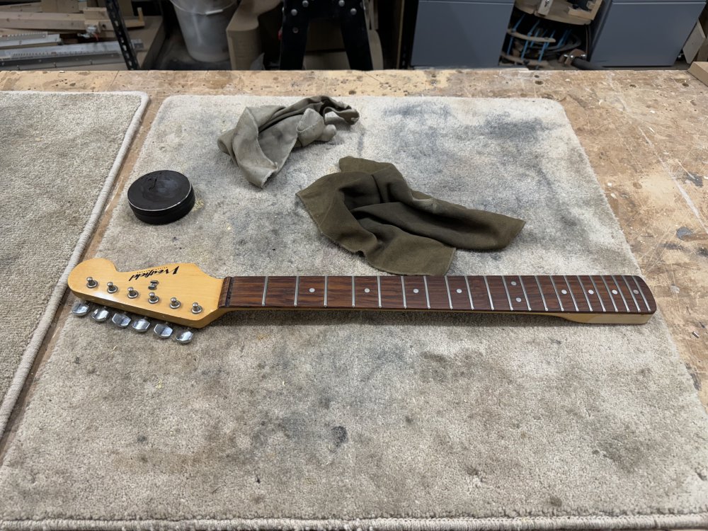 A photo of a guitar neck on a workbench with a start-style headstock. The neck has a rosewood fretboard on maple, and the wood is somewhat at the dark end of rosewood. Next to the neck is what looks like a pot of shoe-polish and some suede cloths.