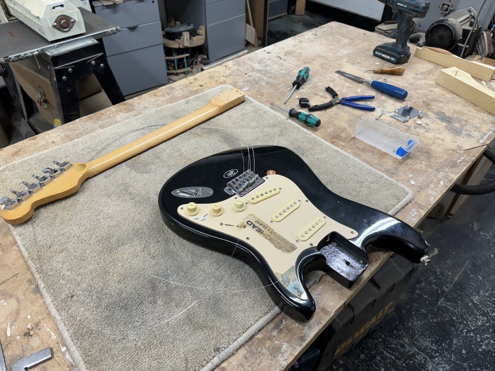 A photo of an electric guitar on a workbench, with the neck disconnected and off to the side, but with the strings still running between them. A strap button can be seen installed on the upper horn of the guitar.