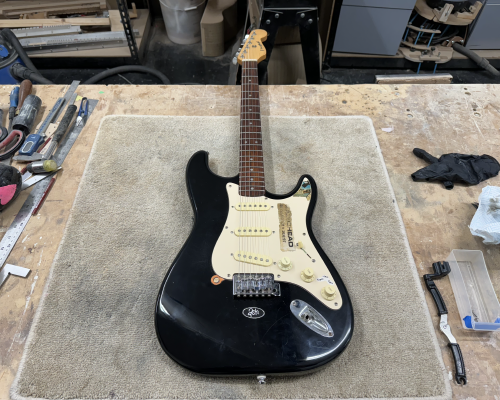 A photo of a black start-clone electric guitar with an off-white pick-guard with stickers on it, and a maple neck with a rosewood fretboard. The guitar is sat on a carpet tile on a workbench in a workshop, with tools to either side of it.