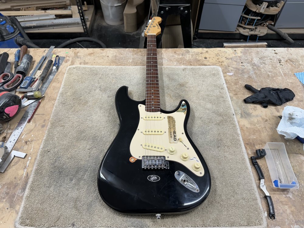 A photo of a black start-clone electric guitar with an off-white pick-guard with stickers on it, and a maple neck with a rosewood fretboard. The guitar is sat on a carpet tile on a workbench in a workshop, with tools to either side of it.