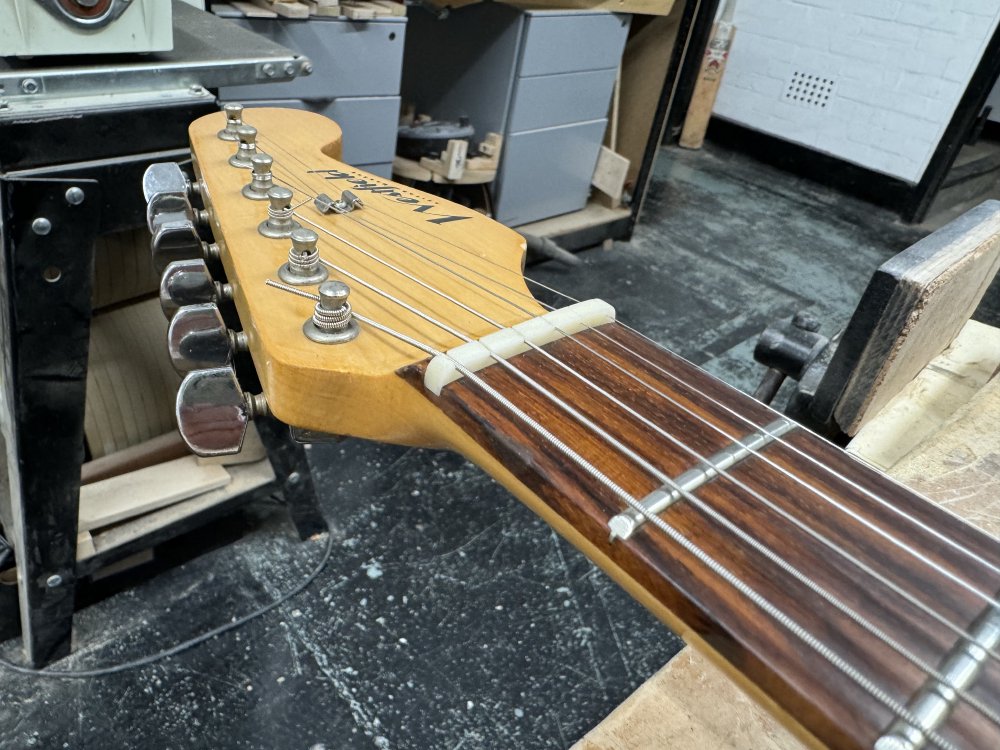 A photo of the top end of the neck of an electric guitar, showing a bone nut with the strings running through it to the tuners.