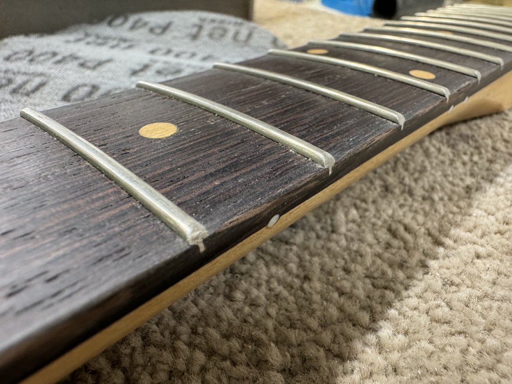 A close-up photo of the edge of the fretboard, showing a few frets where the meet the sides. It's hard to see, but the edge of the fretboard is rounded over between frets, but comes to a distinct line closer to them.