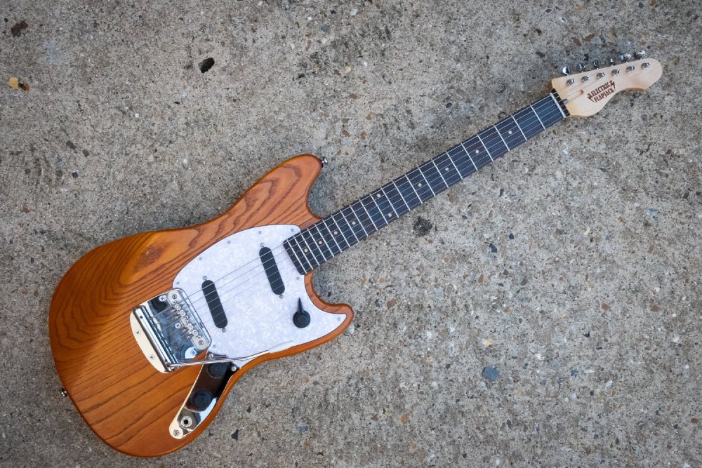 A photo of a solid-body offset style electric guitar laid on a concrete background. The body is natural wood finished, stained orange, it has a white pearlescent pick-guard, into which are set two single coil pickups and a rotary pickup selector switch. It has a tremolo unit in the bridge. The neck is maple with a wenge fretboard.