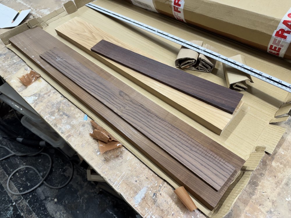 A photo of an opened parcel on a workbench, showing two piles of wood inside. On the left is a long dark coloured piece of roasted-maple on and to the right is a shorter, lighter coloured piece of roasted-maple. Atop each is a piece of rosewood for the fretboard.