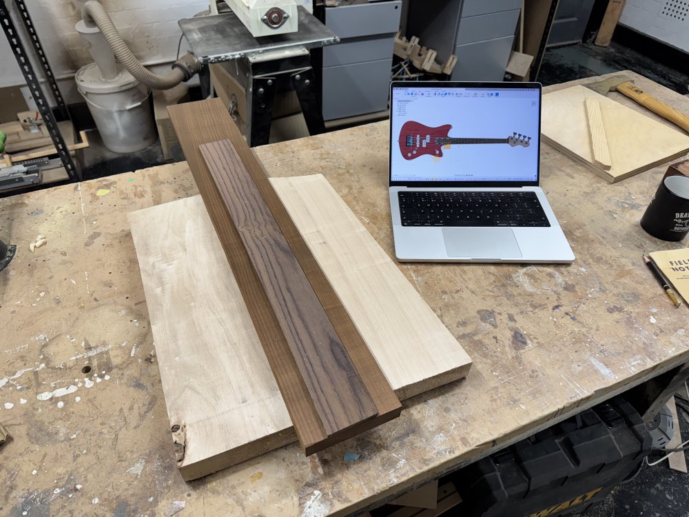 A photo of a piled of three pieces of wood on a workshop workbench: a square of poplar for the guitar body, a plank of roasted maple for the neck, and a piece of rosewood for the fretboard. Next to the wood is a laptop, showing on screen a top down view of a CAD model of a bass guitar.