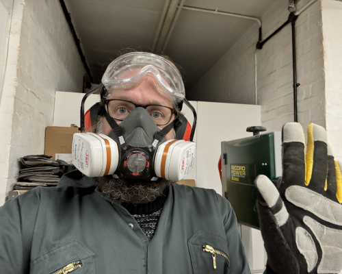 A selfie photo of me in the workshop waving to the camera. I'm wearing a respirator, ear-defenders, eye-protectors, and overalls.