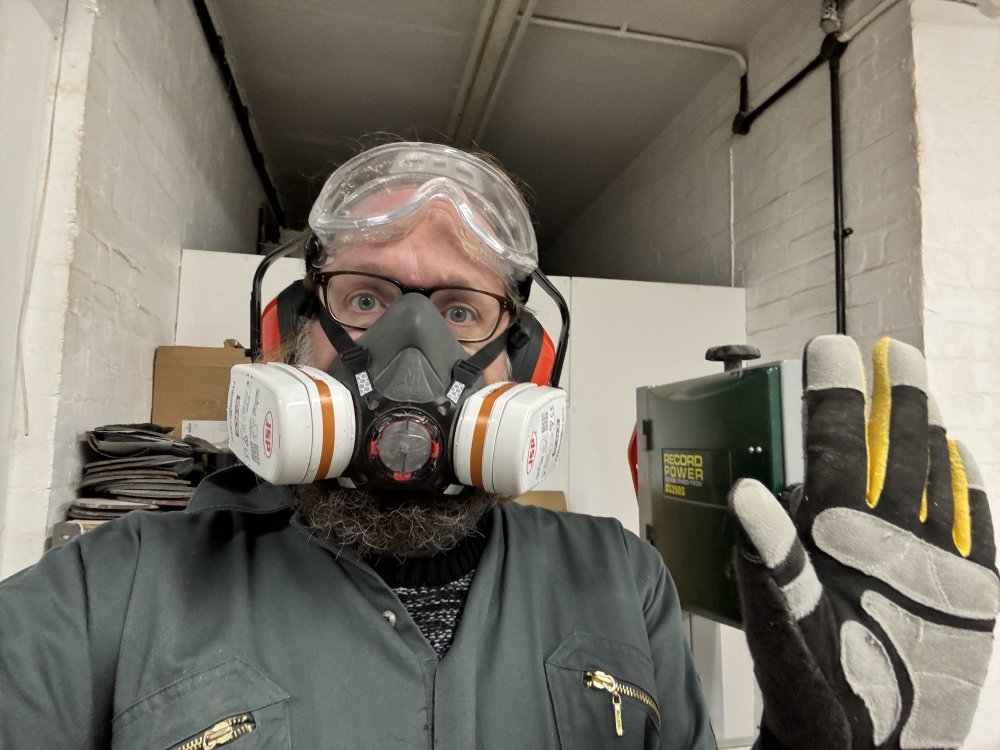 A selfie photo of me in the workshop waving to the camera. I'm wearing a respirator, ear-defenders, eye-protectors, and overalls.