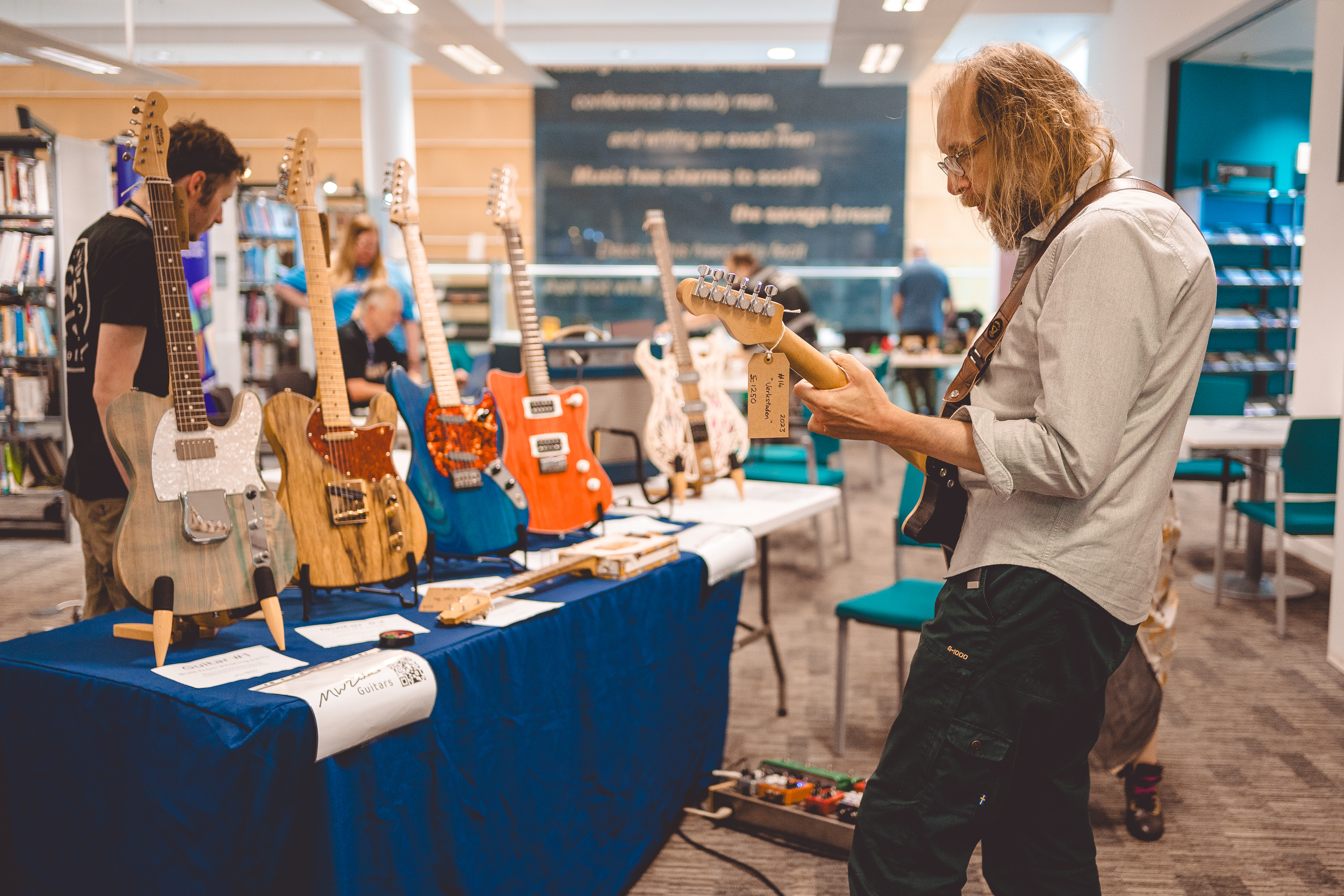 A photo of a man with long hair and a beard stood with his back to the camera playing an electric guitar in a library, stood next to a table that has an array of 5 other electric guitars in different styles.