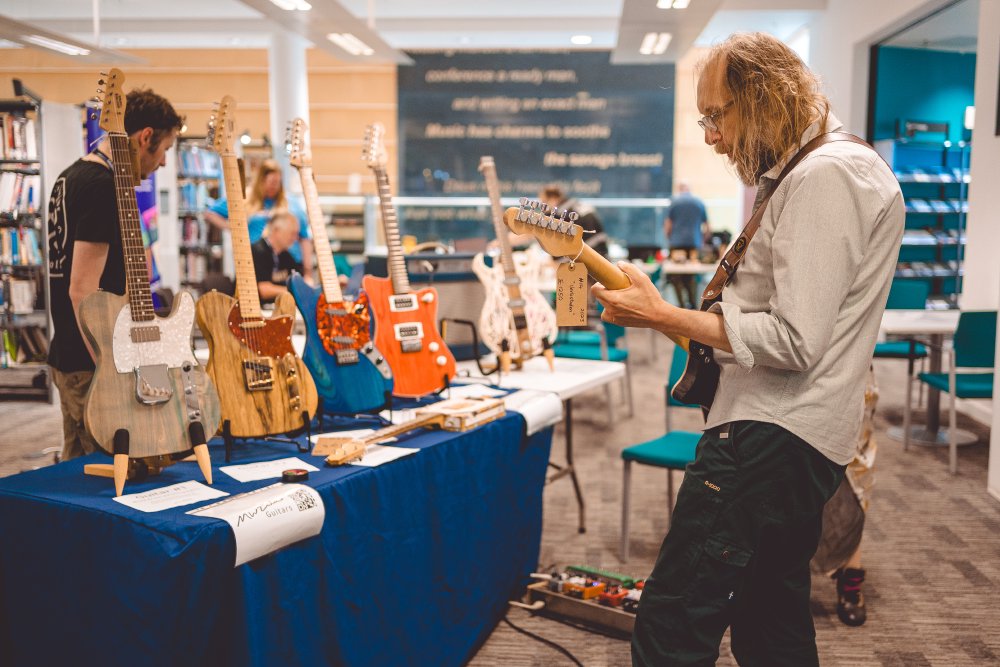 A photo of a man with long hair and a beard stood with his back to the camera playing an electric guitar in a library, stood next to a table that has an array of 5 other electric guitars in different styles.