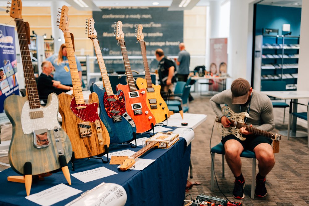 A photo of a person sat in a chair playing an unusual looking electric guitar that is part 3D-printed, part woodwork, next to a table of more conventional looking guitars, all within Liverpool Central Library.