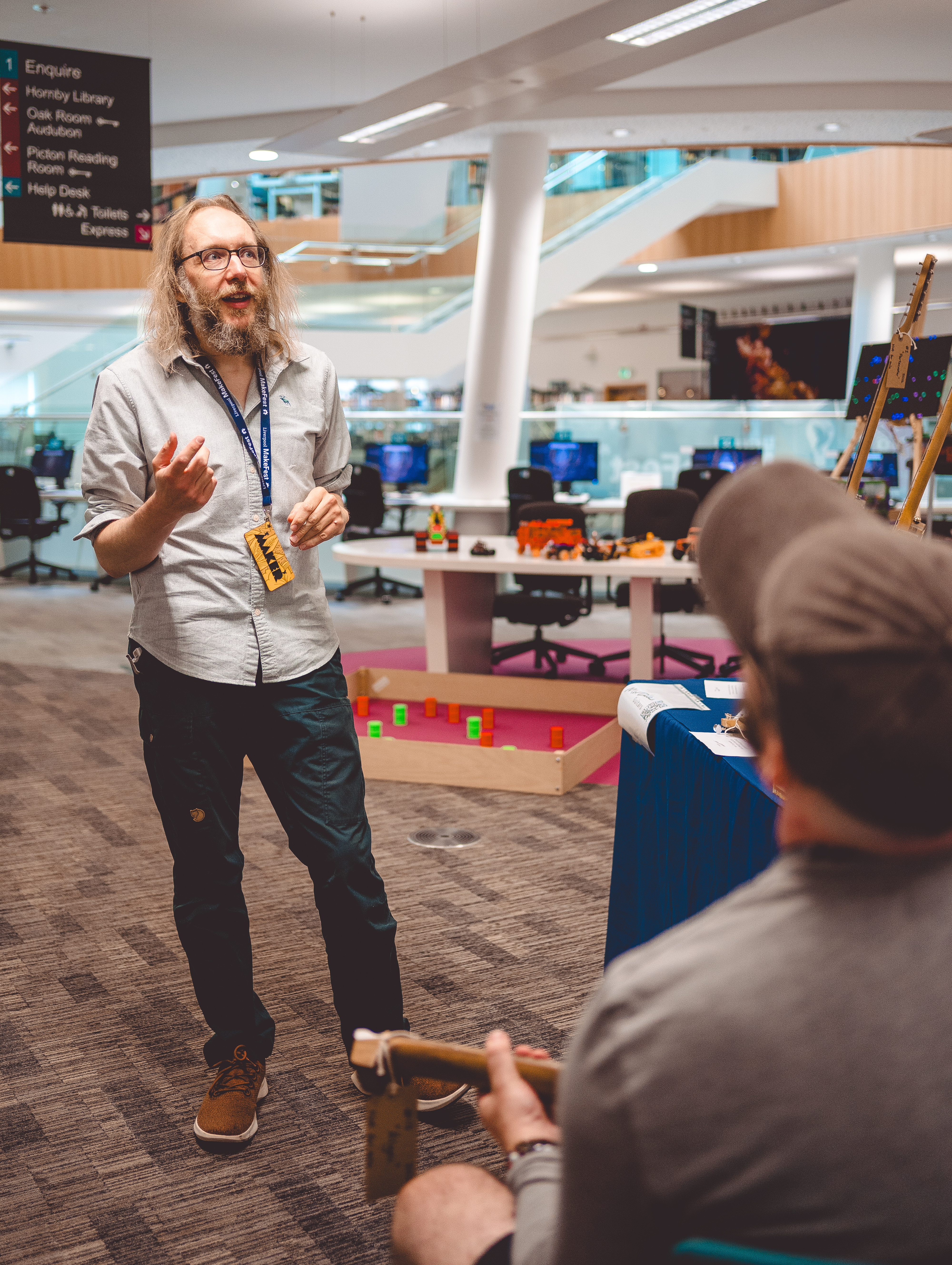 A photo in which a man with long hair and a beard and a badge labelled "maker" is pontificating on something to a person sat down with a guitar.