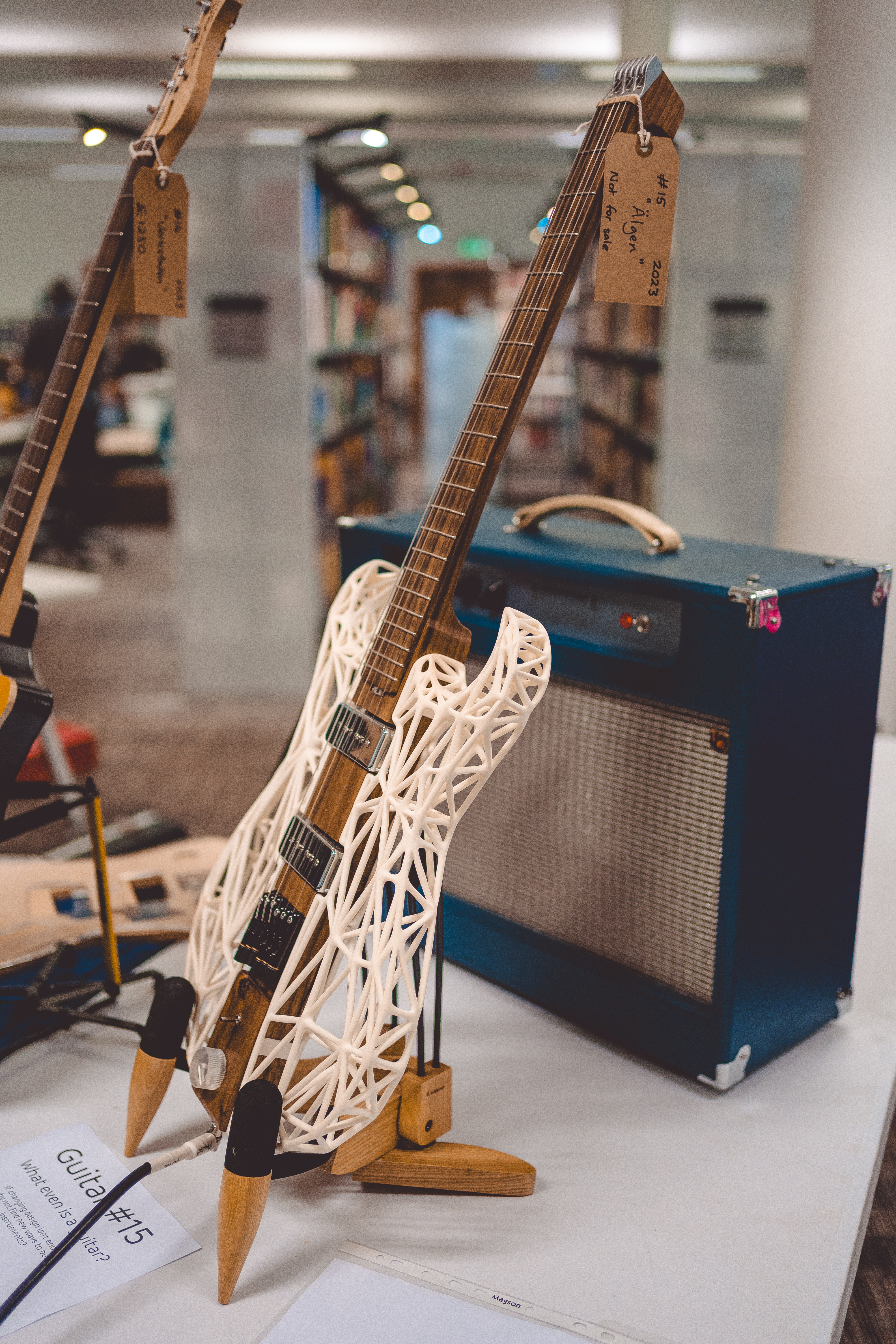 A close up photo of a guitar and an amp on a table in a library. The guitar is an unusual mix of materials, with a central wooden core, but a 3D-printed mesh that makes up the body of the guitar.