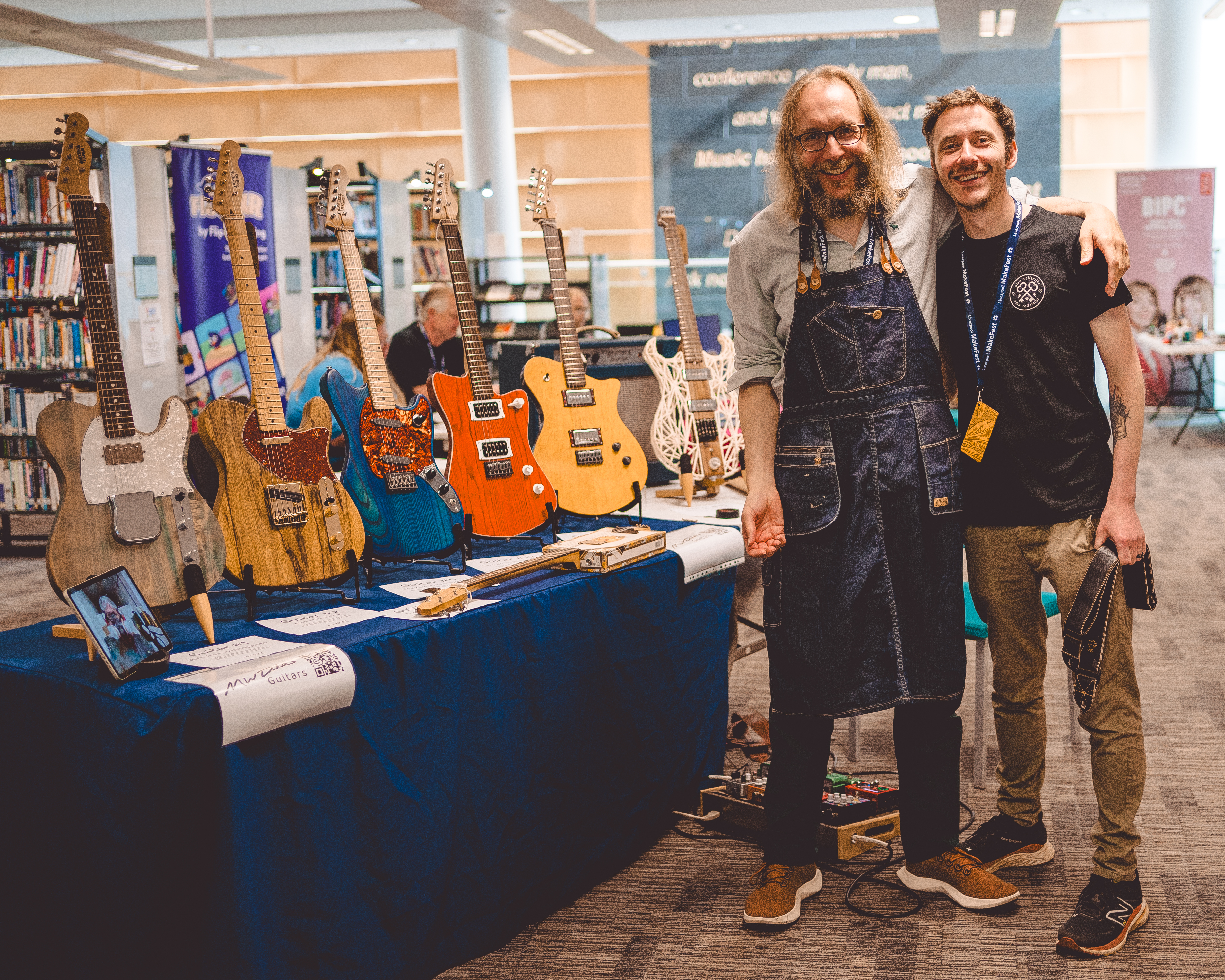 A photo of me and my brother stood together, me with my arm over his shoulder, next to the table full of guitars that we have set up ready for the Makefest crowd to come see.