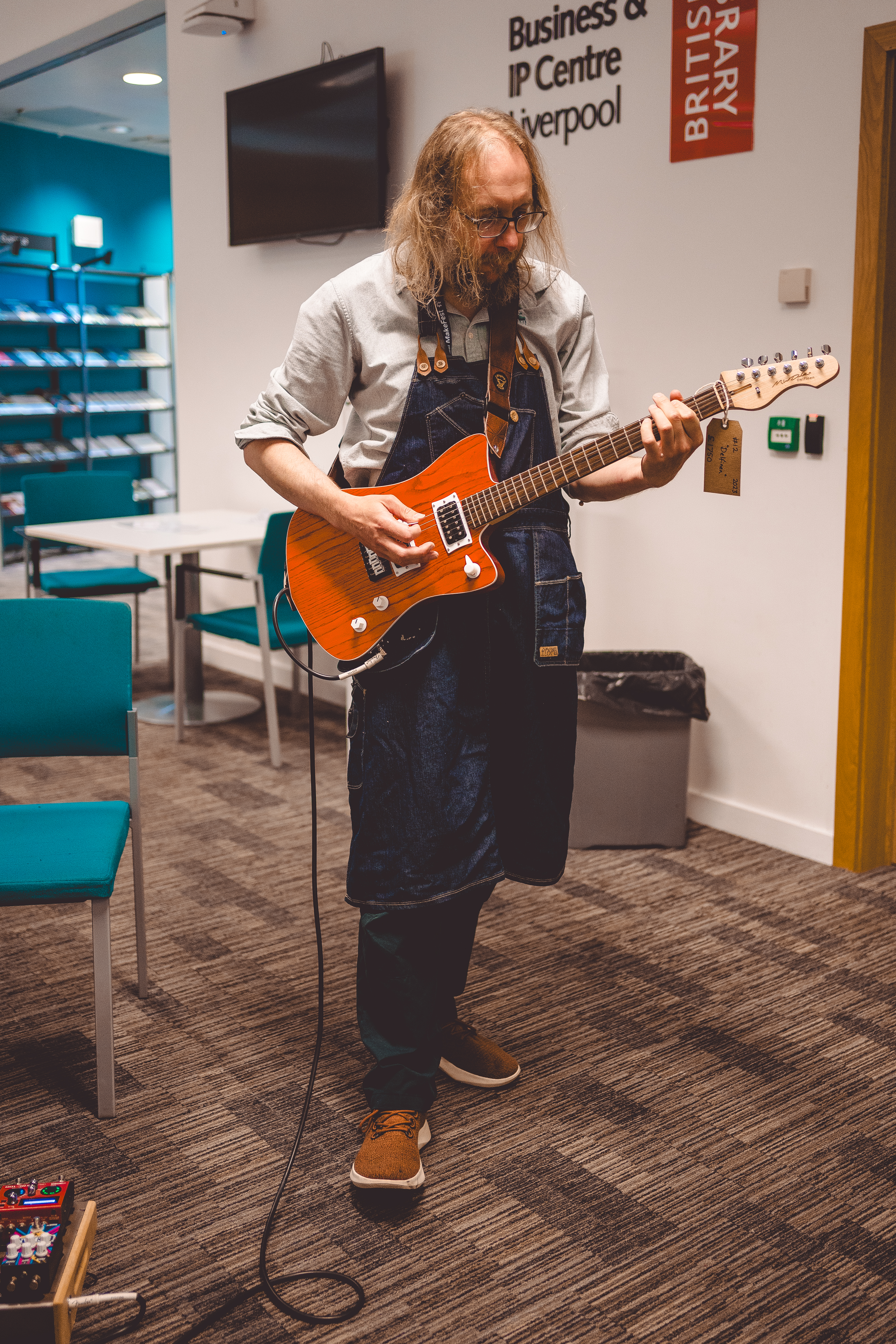 A photo of me in Makefest garb, which is regular clothes with a denim workshop apron over them, playing an orange finish offset electic guitar whilst stood in a library.