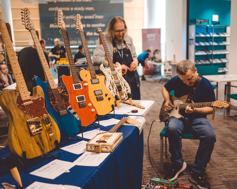 A photo of a man sat playing a faded-blue telecaster style guitar, next to a table with a bunch of other guitars on it in different shapes and colours. In the background I'm stood watching the man play with a smile on my face.
