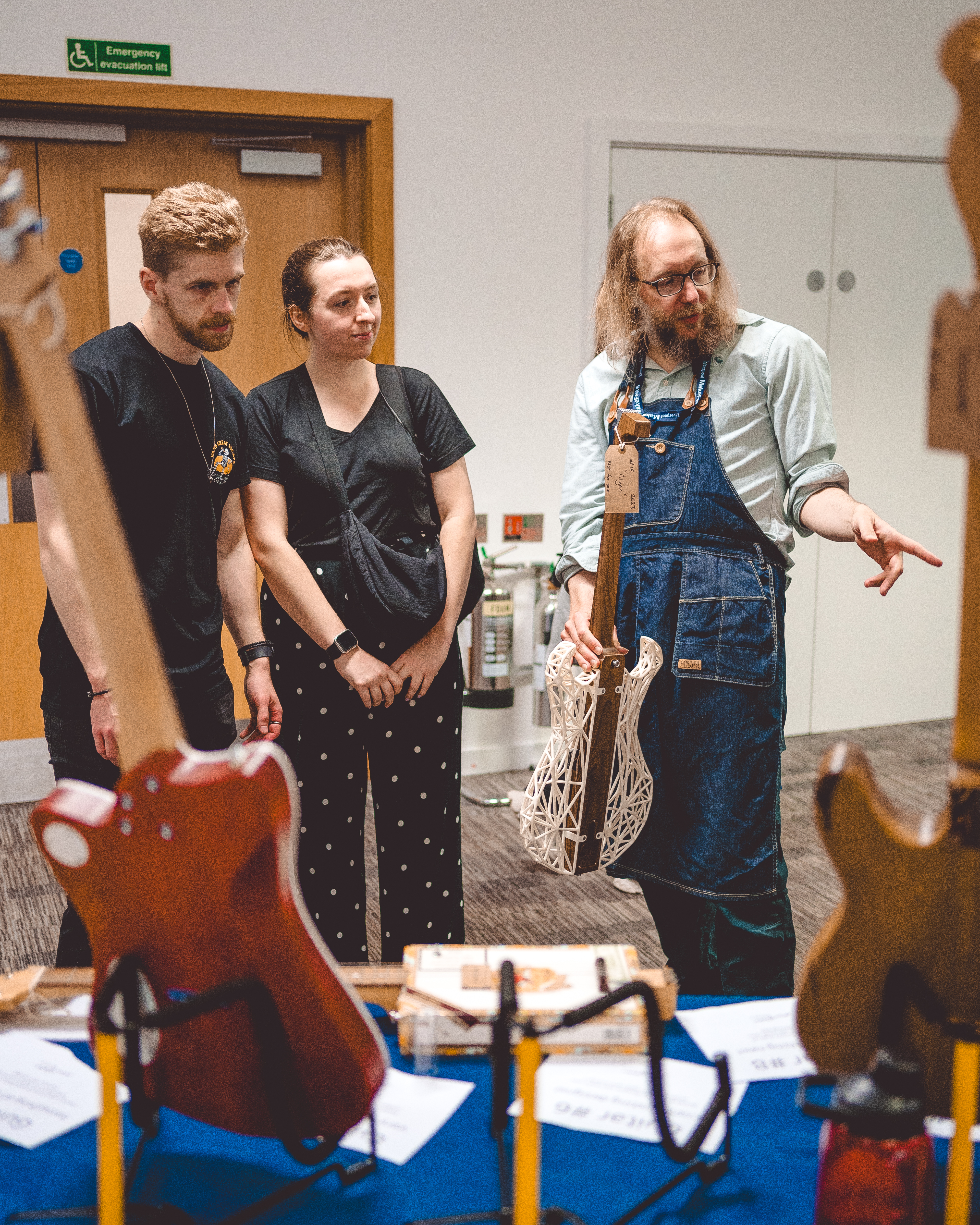 A photo of me chatting two two people at Makefest. In one hand I'm holding the partially 3D-printed guitar, and with the other I'm gesturing to one of the other guitars on the table. The other two people are looking with some interest at whatever I'm gesturing to.