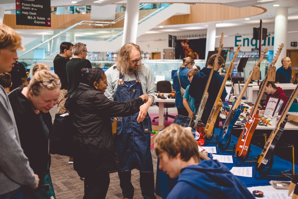 A photo of a busy library of people, with various other makers and their tables in the background, and my table with 5 guitars stood on it in the foreground. I'm stood chatting to a lady who is gesturing to one of the instruments.