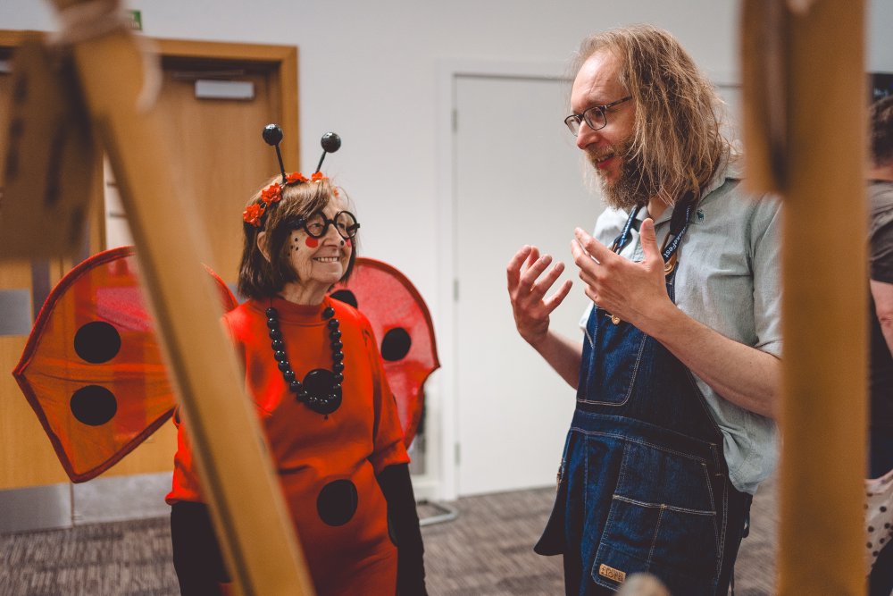 A photo of me stood next to some guitars whilst wearing a workshop apron over my regular garb, stood chatting explaining something to a lady dressed and made-up like a ladybird.