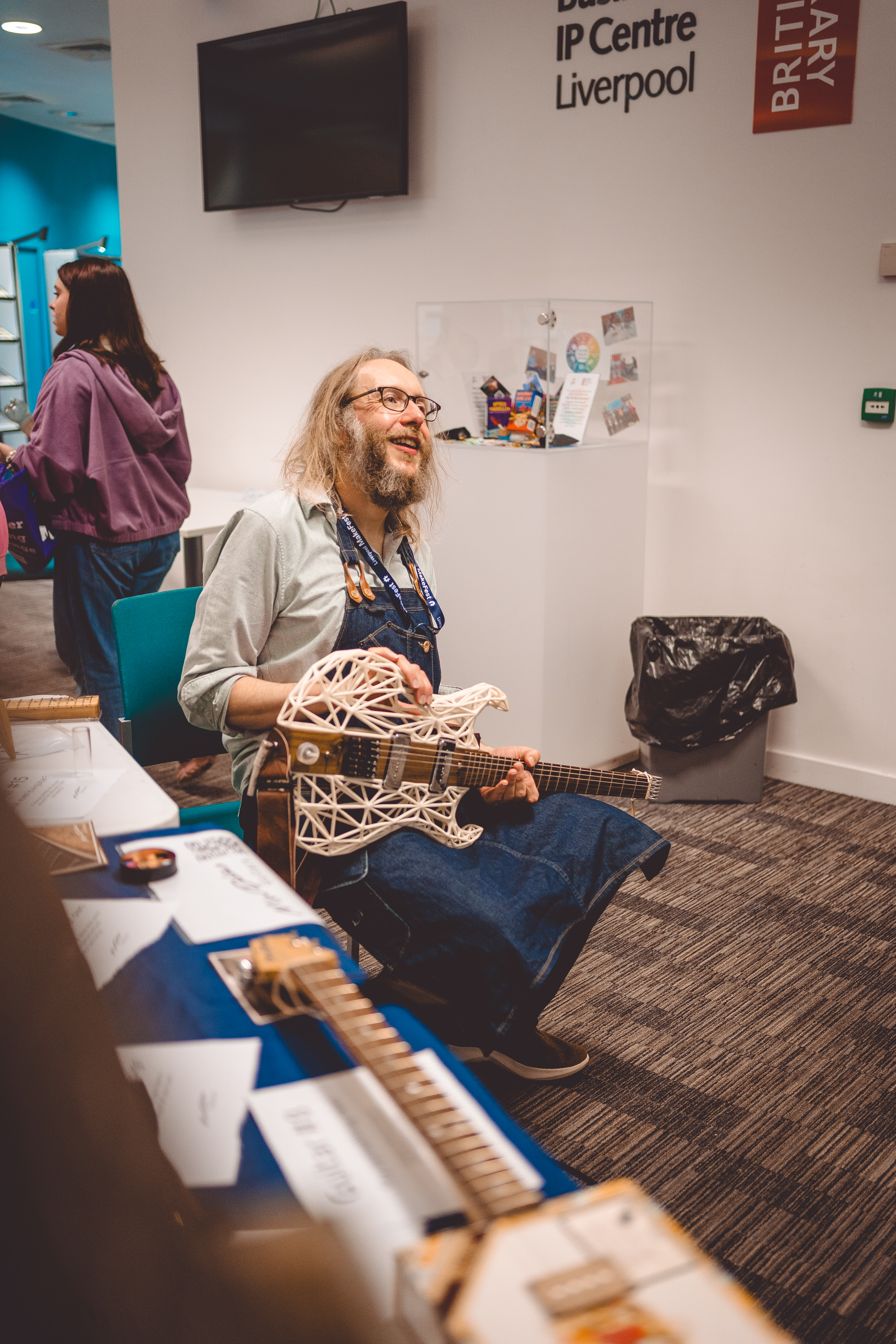 A candid photo of me sat on a chair, holding the wooded/3D-printed guitar on my lap but not playing it, and I'm looking up as if in coversation with someone and I have a smile on my face.