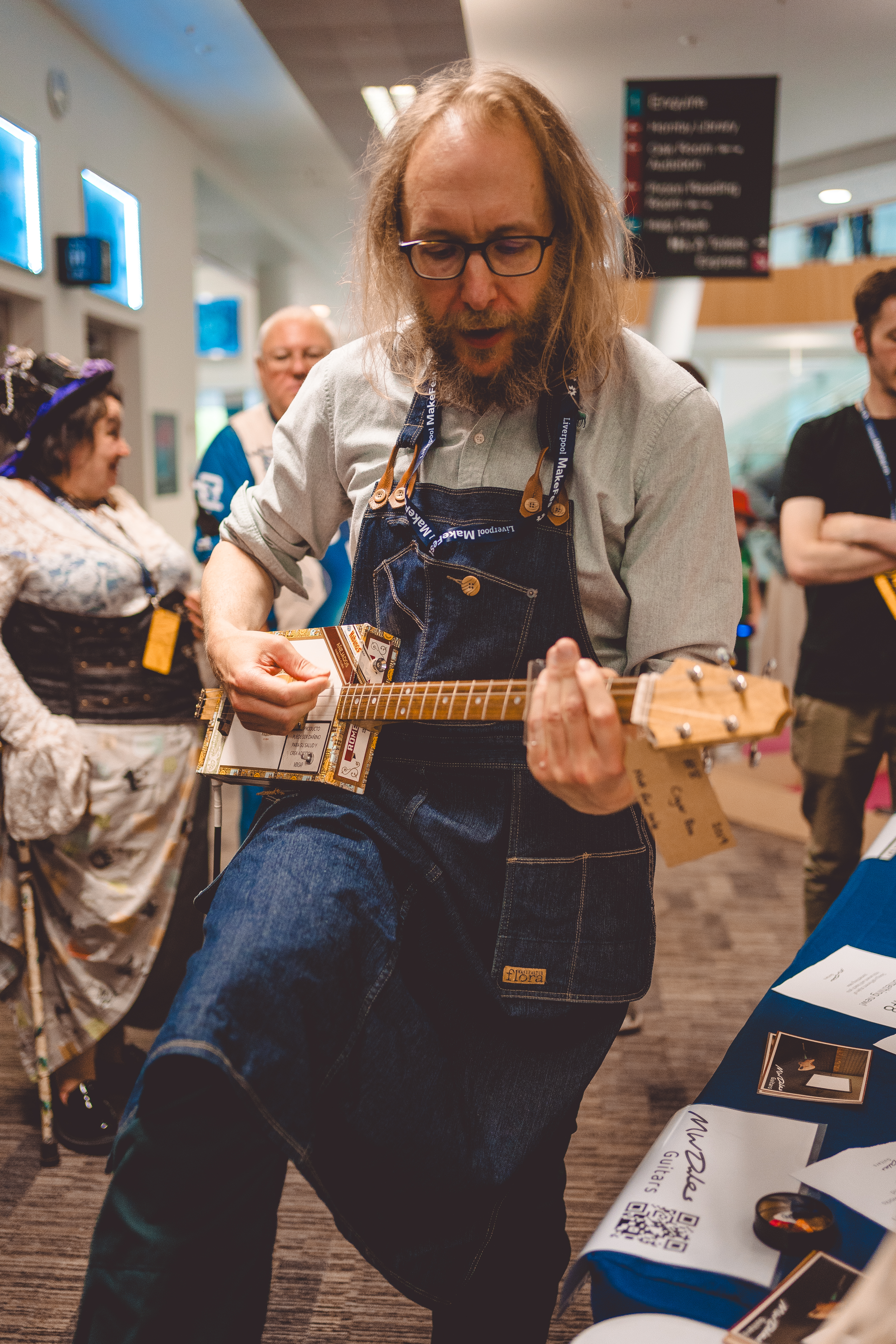 A close up photo of me, with a collection of people in the background, attempting to play slide guitar on the cigar box guitar.