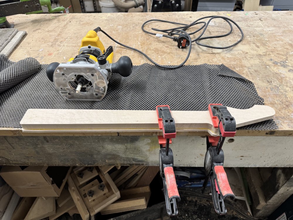 A photo of a work in progress guitar neck clamped to a workbench, next to which sits a De Walt palm router.