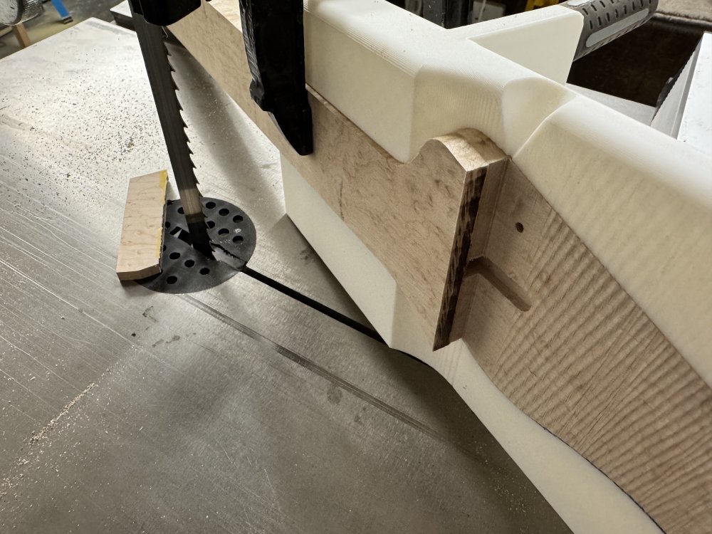 A close up photo showing a step down from the fretboard to the headstock, with the neck in the 3d-printed jig, all of which is sat on a band-saw table.