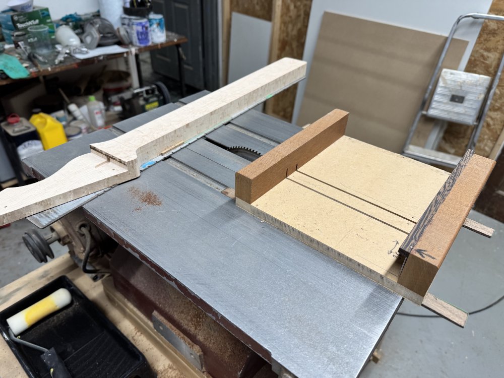 A photo of a small table-saw bed, and on one end sits a guitar neck being built, sat on a metal plate with regular notches in the side, and at the other side is a wooden sled that runs parallel to the cutting blade.