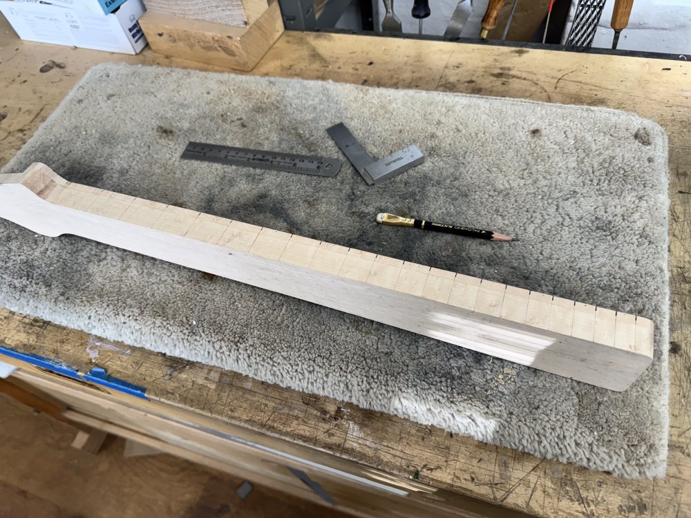 A photo of the work-in-progress guitar neck sat on its side on a workbench, showing the side that the player would look down into whilst playing. Where the fret slots have been cut, a pencil line has been drawn extending the line down the side of the neck, clearly indicating fret positions on the side.