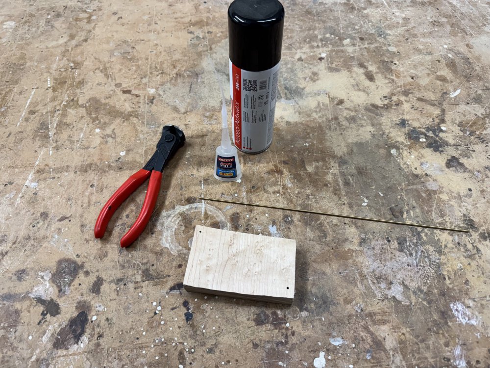 A photo of a rectangular scrap of maple on the workbench with a couple of small (2mm-ish) holes in it. Next to it is a thin dowel (also 2mm or so diameter) of golden metal, a pair of metal cutting pliers, and some superglue.