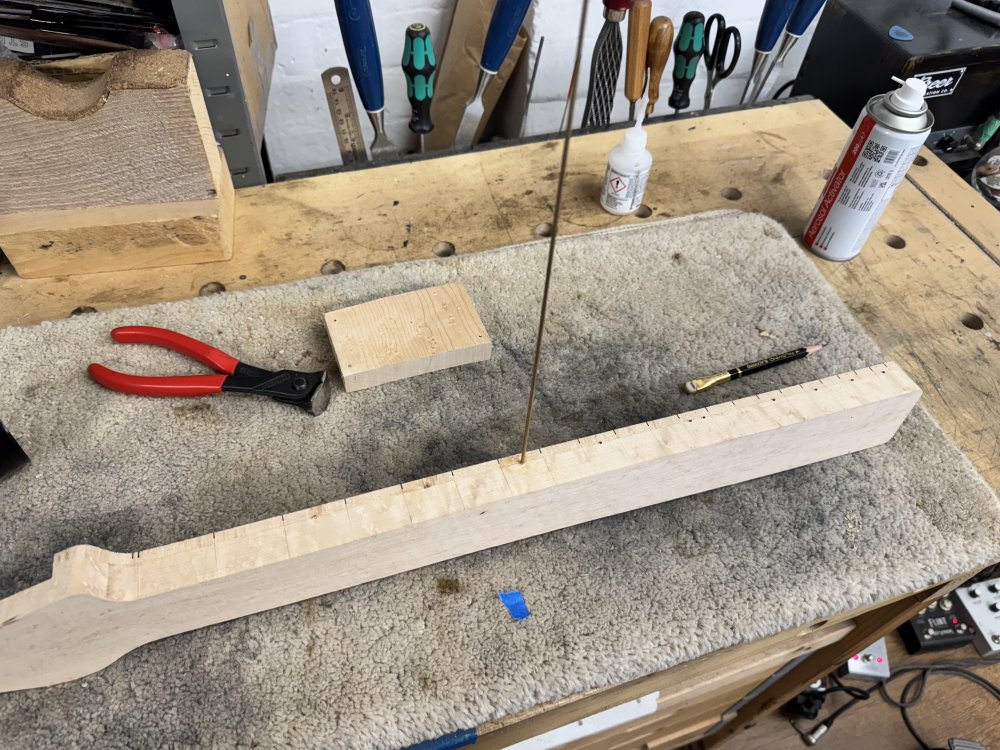 A photo of the work in progress neck, on its side on the workbench, and with the 20cm long dowel of goldenish metal sticking out of one of the holes drilled in the side of the neck for the inlays.