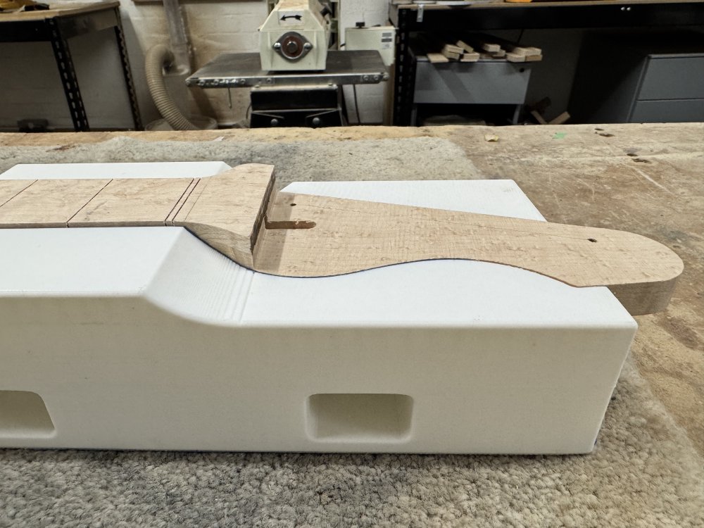 A close up photo again of the stepped transition from the fretboard to the headstock level on the neck. It sits in the 3D-printed jig, and you can see that the jig has a curve behind where the step is, indicating what the neck should look like once the step has been removed.