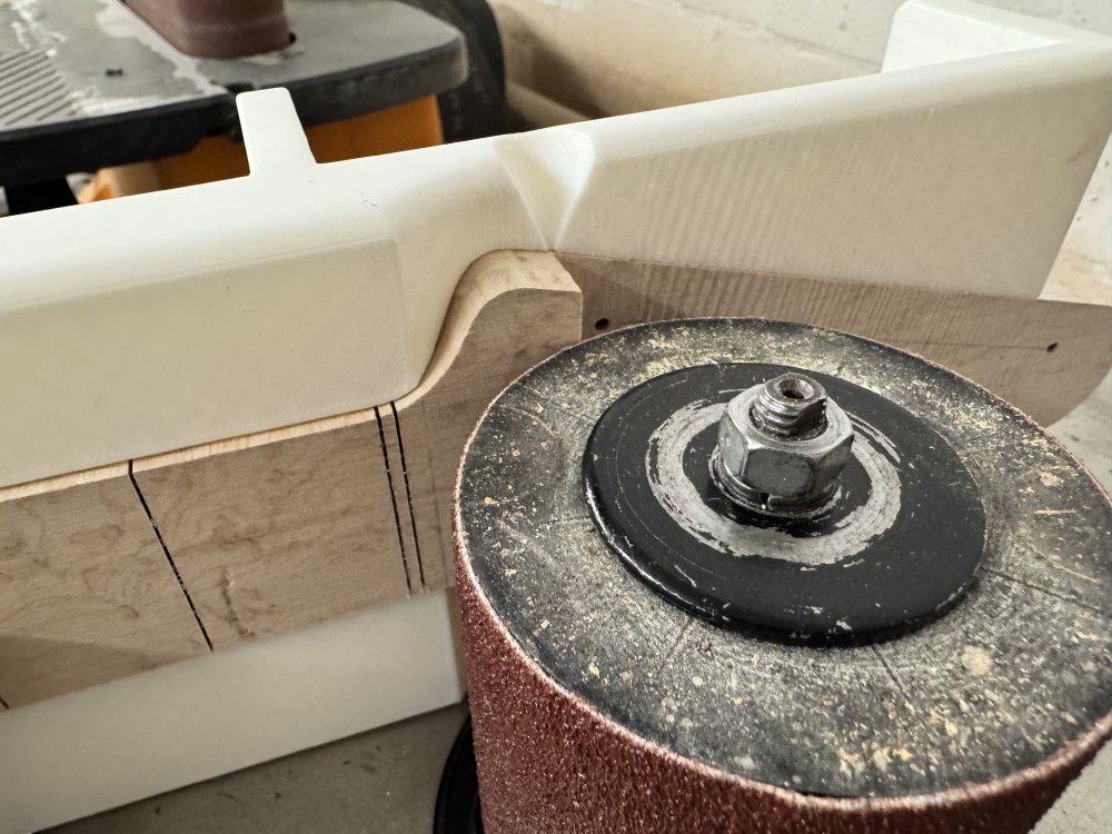 A photo of the neck in the jig sit on the table of a spindle sander, with the step pressed up against the spindle ready for sanding.