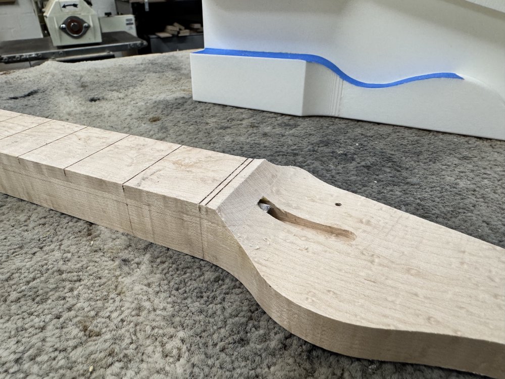 A photo of the work in progress neck sitting on a workbench, and where before there was a step from the fretboard level to teh headstock level, there is now a nice rounded transition.