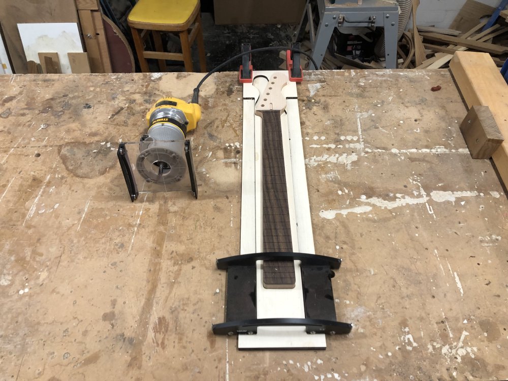 A photo of a guitar neck sat in a plywood jig that runs the length of the neck and guides a metal arch up and down the length of the fretboard. The arch will support the palm-router that is sat to one side.