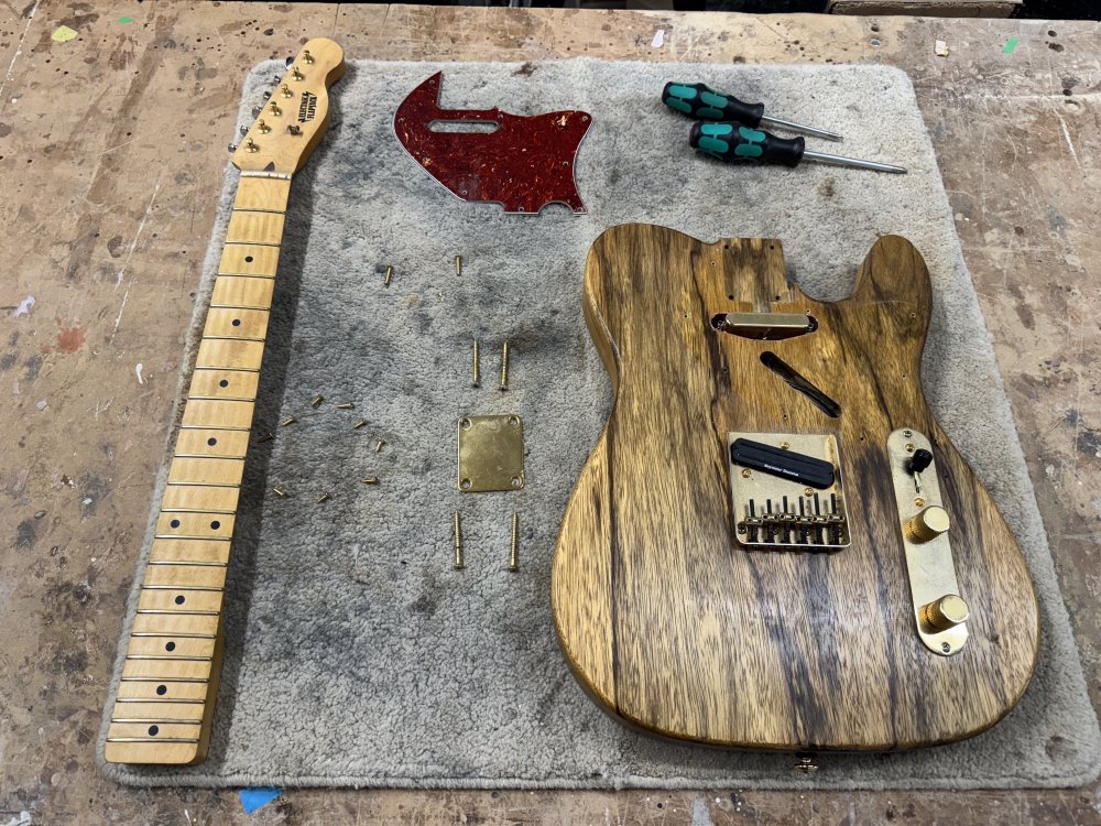 A photo of the electric guitar from the first picture, showing it now disassembled into parts on the workbench: the neck to one side, the body to the other, the pick-guard in the middle and screws and screwdrivers around it.