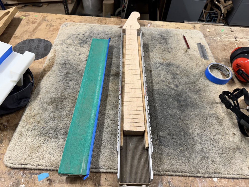 A photo of a work-in-progress electric guitar neck sat in a metal frame that has parallel rails up the length of either side of the neck. To one side is a metal beam with sandpaper stuck to it that has a slight concaved radius to it.