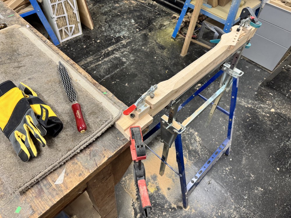 A photo of the work-in-progress guitar neck clamped onto a trestle stand, with wood dust on the floor. The neck is not yet properly carved, still looking plank like, but into the sides of the rear of the neck are two facets at a close to 45 degree angle, running from the first fret down to around the 15th fret. Next to the neck sits a very brutal looking saw-toothed rasp.