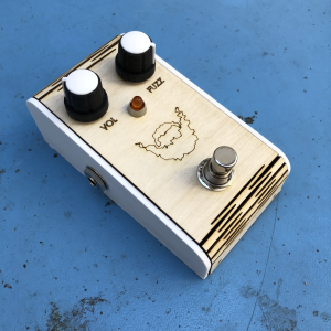 A guitar effects pedal sits on a workbench. It has two control knobs, labelled VOL and FUZZ, a footswitch, and a picture of fuzzy beard outlined on it. The lower case is 3D printed white ABS, the top surface us laser etched plywood with a living hinge letting it wrap around the top and bottom edges.