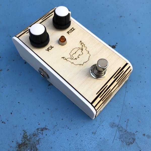 A guitar effects pedal sits on a workbench. It has two control knobs, labelled VOL and FUZZ, a footswitch, and a picture of fuzzy beard outlined on it. The lower case is 3D printed white ABS, the top surface us laser etched plywood with a living hinge letting it wrap around the top and bottom edges.