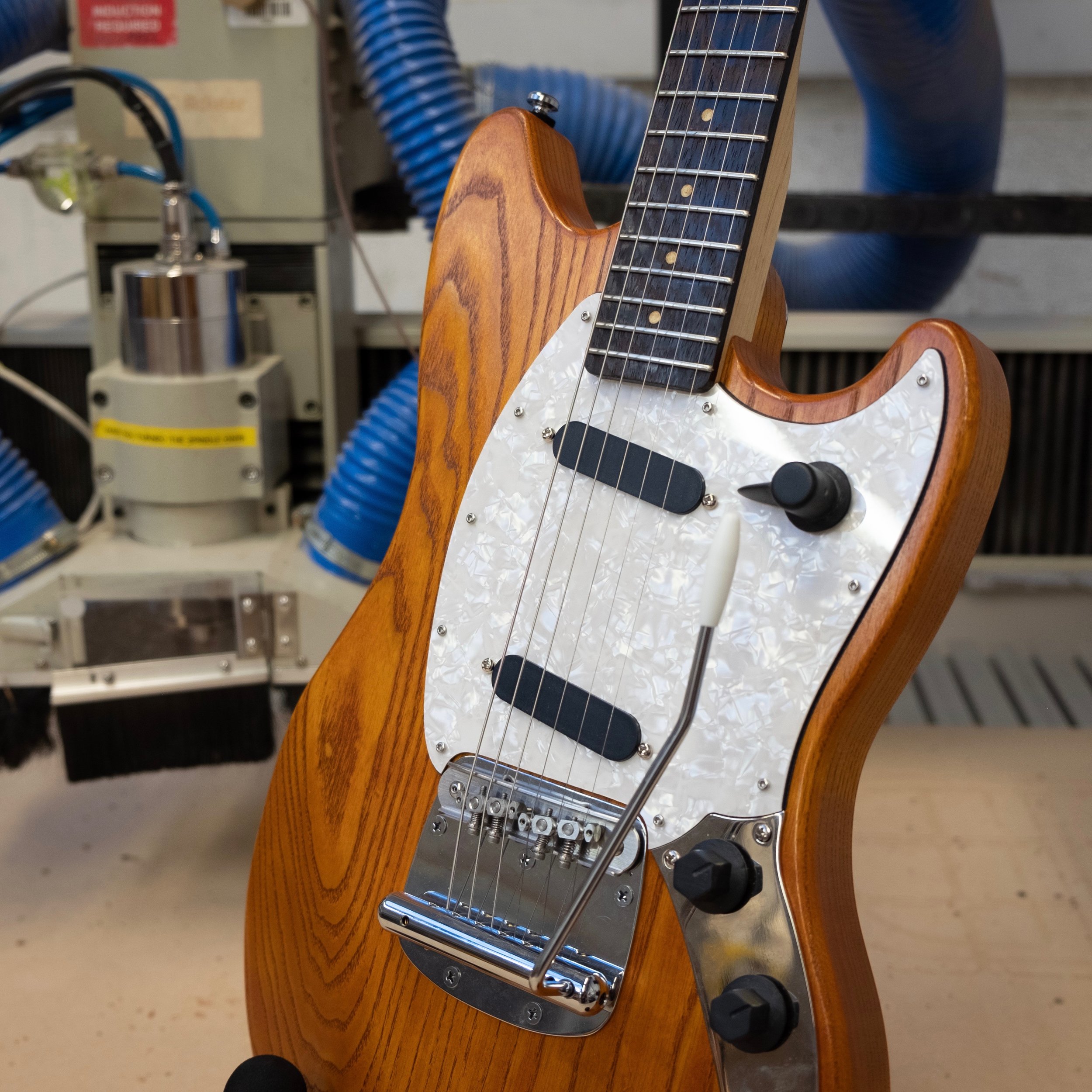 A close up of the body of a mustang-style guitar, showing the orange body and white pearloid pickguard, black single-coil pickups, and custom 3D-printed black control knobs.