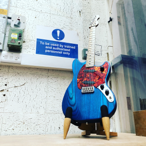 A blue mustang style guitar with a marbled red pickguard and a maple nect sits in a stand in the workshop, next to a workshop sign that reads 'To be used by trained and authorized personnel only.'