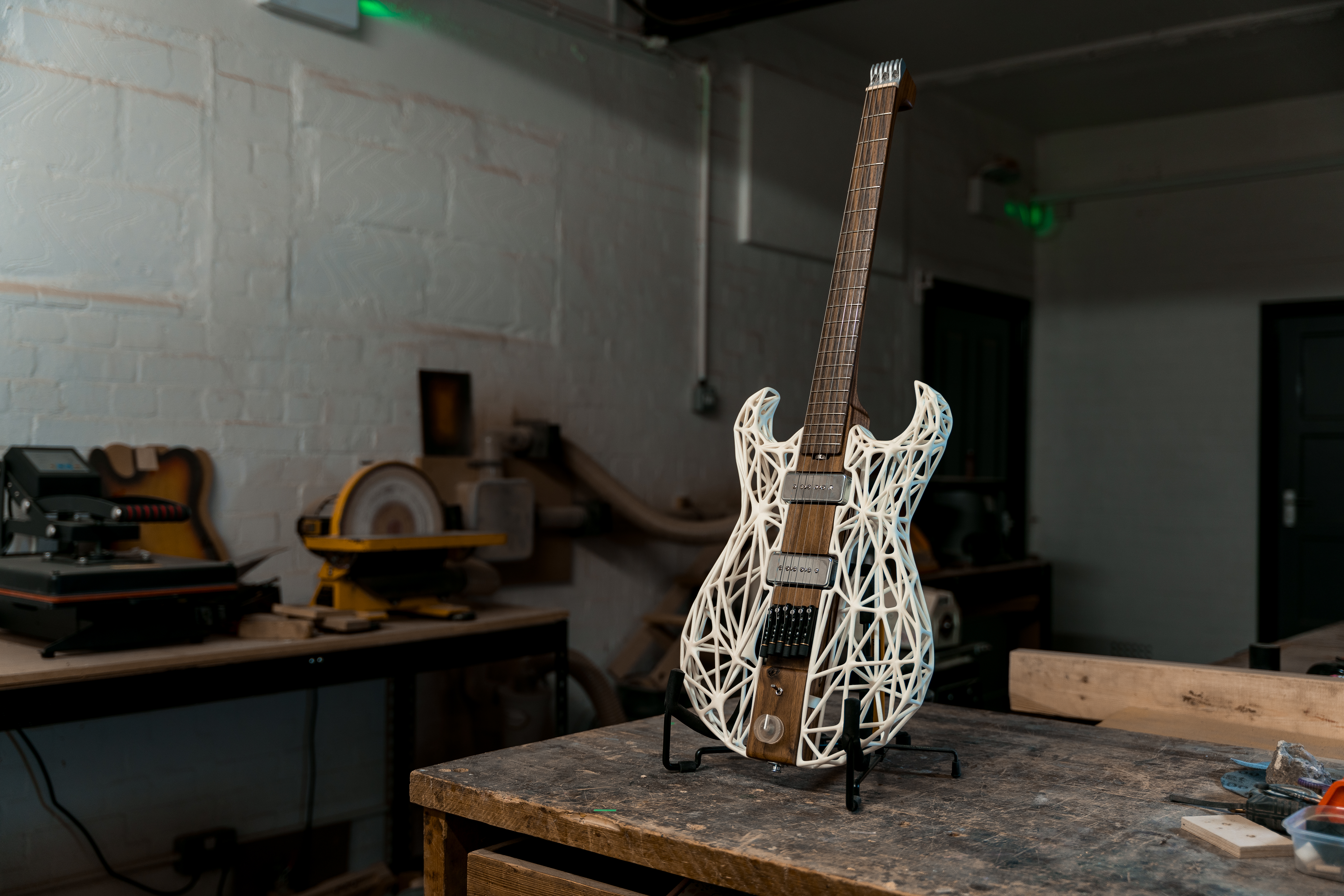 A photo of an unusual looking guitar siting on a stand on a workbench in a moodily lit workshop. The neck and the centre of the body along the neck are made from a single piece of dark walnut wood, whilst the sides of the body are made from a white 3D-printed lattice work. There is no headstock, just a custom aluminium block to anchor the strings, with tuners being built into the bridge.